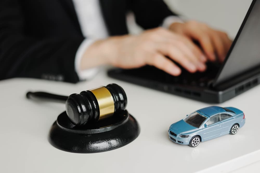 Gavel and toy car on desk with person typing on laptop in background.