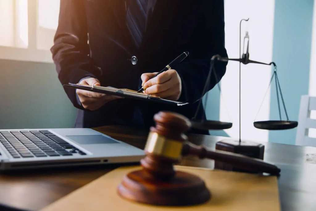 Lawyer reviewing legal documents on clipboard with gavel, scales of justice, and laptop on desk in law office. Lawyer reviewing legal documents on clipboard with gavel, scales of justice, and laptop on desk in law office.