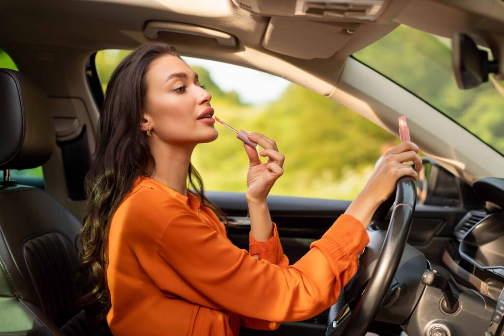Woman applying lipstick while driving car, illustrating distracted driving and unsafe road behavior.