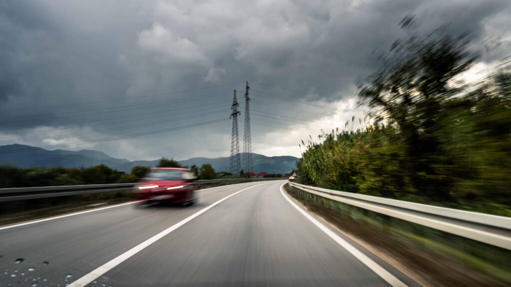 Red car speeding on wet highway under dark storm clouds, representing dangerous driving and poor weather road conditions.